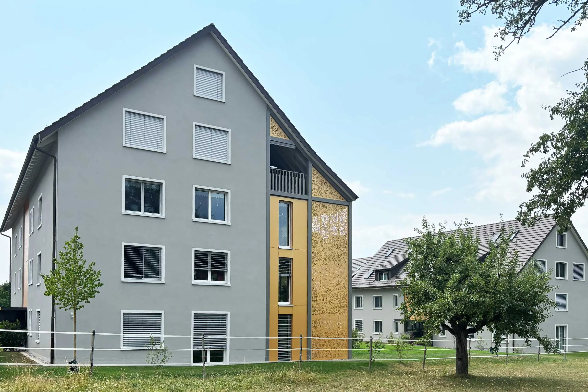 Ansicht Gibelseiten von Mehrfamilienhaus mit grauer Fassade und orangener Metallverkleidung am Rand