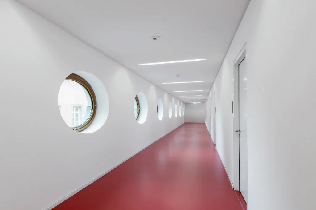 Weiss gestrichener langer Flur mit Bullaugenfenster und rotem Bodenbelag 